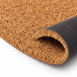 A close-up of a brown coir doormat with a black textured underside, shown partially rolled to reveal the backing on a white background.