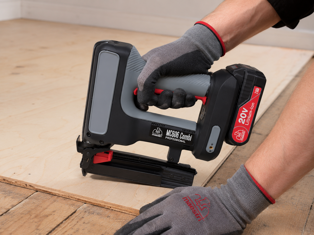 A cordless nail gun being used by a person wearing gray and red work gloves on a light wood sheet, with a plywood board and wooden floor visible in the background.