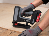 A cordless nail gun being used by a person wearing gray and red work gloves on a light wood sheet, with a plywood board and wooden floor visible in the background.