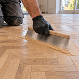 Gloved flooring professional spreading smooth wood filler across sanded oak parquet with a wide flat-bladed filling trowel.
